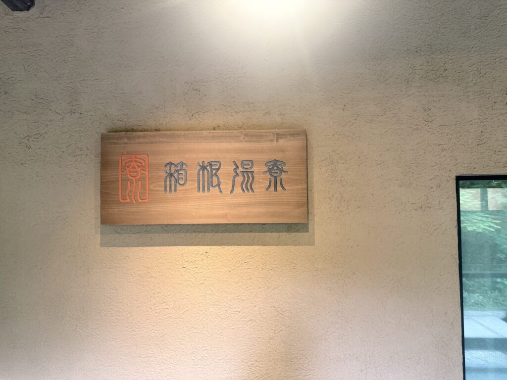 「箱根湯寮」と力強く彫られた木製の壁掛け看板。柔らかい照明に照らされ、高級感と落ち着きのあるUIデザインを体感させる一枚。